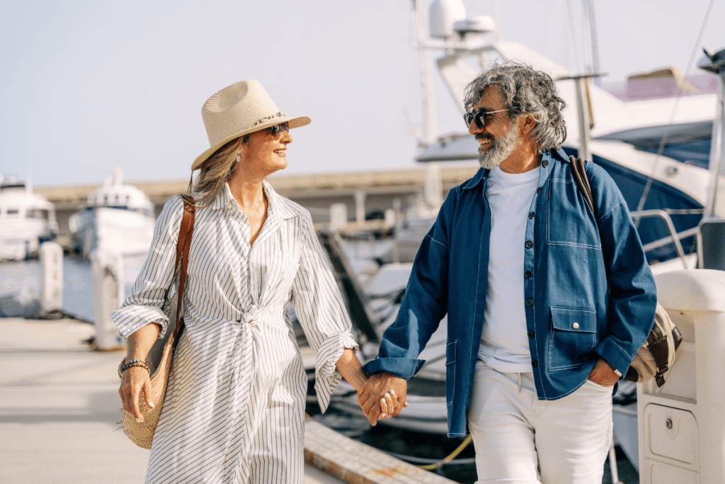 Senior couple holding hands, smiling, and walking by upscale boats in a sunny marina.