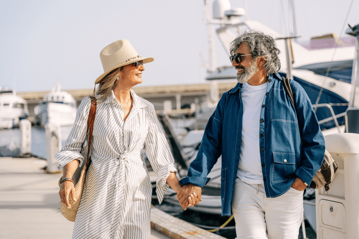 Senior couple holding hands, smiling, and walking by upscale boats in a sunny marina.
