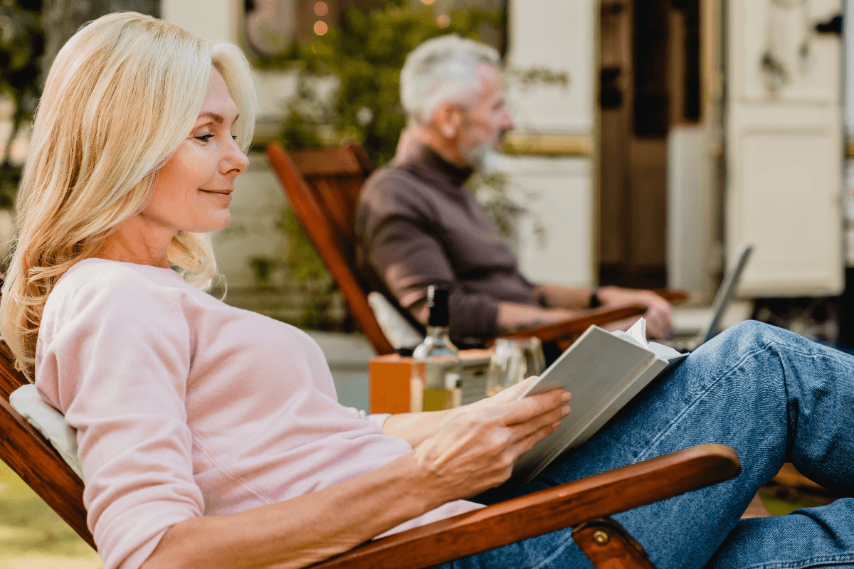 Content senior blond lady reading a book in the foreground with her grey-haired husband on the background in the garden with their trailer behind.